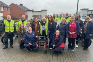 A group of individuals is gathered outdoors on a paved surface in front of a metal fence and several parked motorcycles. One individual stands at the front using a mobility aid and wearing a high‑visibility vest. The rest of the group is positioned around and behind this person, with some individuals wearing high‑visibility jackets and others wearing outdoor clothing or uniforms. Residential buildings and a cloudy sky are visible in the background.