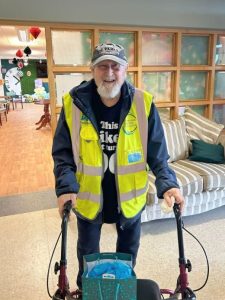 An individual stands indoors using a mobility aid. The individual is wearing a high‑visibility vest over a dark top and a cap with visible printed text that includes the number “1936.” The person is positioned in the foreground, holding the handles of the mobility aid. A gift bag is placed on the seat section of the mobility aid. Behind the individual, there is a sofa with striped upholstery, cushions, and a wooden wall feature with glass panels. Further into the background, more furnishings and decorations are visible, including balloons and hanging décor in a room with a wooden floor.