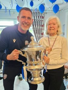 A Coleraine FC representative wearing a navy tracksuit stands beside a Ratheane Care Home resident as both hold the silver Irish Cup trophy with blue and white ribbons. The room is decorated with blue and white balloons hanging from the ceiling and patterned curtains in the background.