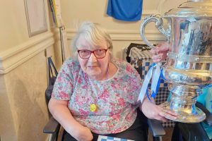 A Ratheane Care Home resident seated in an armchair beside the Irish Cup trophy, which is being held at the side. The resident is wearing a floral top and a medal on a red, white and blue ribbon, with Coleraine-themed decorations and a blue flag visible on the wall behind.