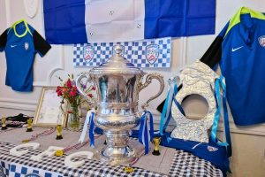 The Irish Cup trophy displayed on a table decorated with blue and white Coleraine FC colours, including flags, scarves and jerseys. The display also includes small gold trophies, medals, flowers in a vase, and a silver decorative piece arranged around the cup.