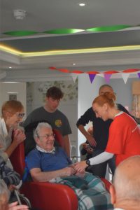 Residents, visitors and volunteers gathered at Milesian Manor Sports Day, with one resident seated in a chair while taking part in an activity surrounded by supporters and colourful decorations.
