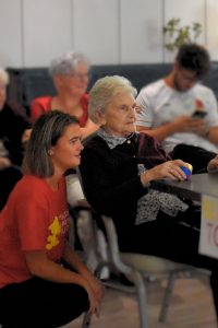 Resident taking part in a Sports Day activity at Milesian Manor, seated at a table holding a small ball while a Children's Heartbeat Trust volunteer sits nearby and other attendees watch in the background.