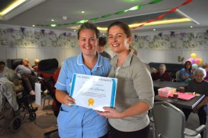 Two members of staff hold a recognition certificate during Milesian Manor's Sports Day fundraiser, with residents seated in the background and colourful bunting and streamers decorating the room.