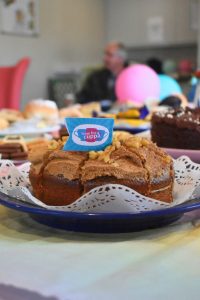 Close-up photograph of a homemade cake displayed on a blue serving plate lined with a decorative paper doily at a fundraising coffee morning. The cake is topped with a cinnamon-coloured frosting and chopped nuts, with a small sign reading “Time for a Cuppa” placed in the centre. In the softly blurred background, additional cakes, traybakes and refreshments can be seen on a decorated table, alongside colourful pink and turquoise balloons and guests attending the event.