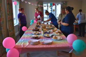 A long table covered with a colourful pastel tablecloth is filled with homemade cakes, traybakes, scones, pastries and other baked goods at a fundraising coffee morning. The room is decorated with pink and turquoise balloons and bunting, while several residents, visitors and care home staff gather around the table selecting refreshments and chatting. Large windows and patterned curtains brighten the room, creating a welcoming community atmosphere.