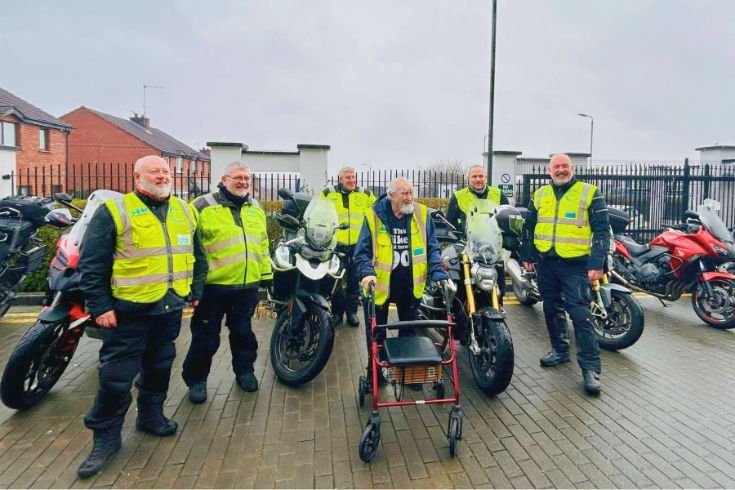 A group of individuals is gathered outdoors on a paved area in front of a metal fence with residential buildings visible in the background. Several motorcycles are parked behind the group. One individual is positioned at the front using a mobility aid, wearing a high‑visibility vest. Other individuals around them are wearing a mix of high‑visibility clothing, outdoor jackets, and uniforms. The group is arranged closely together, facing the camera.